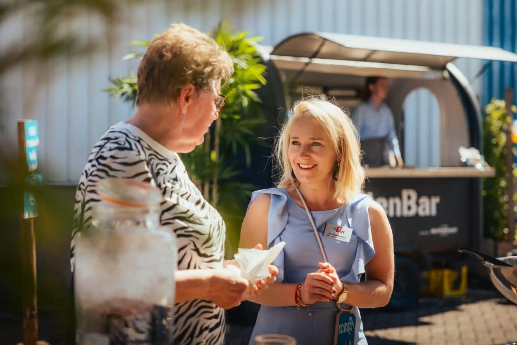 Twee vrouwen praten bij een buitenkraam.